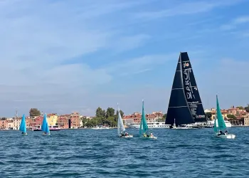 Piccole derive Hansa con vele azzurre e bianca davanti a un grande maxi yacht dalla vela nera, sullo sfondo gli edifici di Venezia
