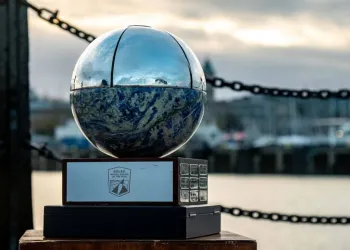 Trofeo a forma di sfera in pietra blu e metallo lucido su base in legno, con catene e specchio d'acqua sfocati sullo sfondo