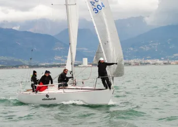 Barca a vela J24 con quattro persone a bordo naviga con spinnaker su mare mosso, con montagne e costa sullo sfondo