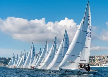 Fila di barche a vela J/22 in regata davanti al lungomare di Napoli con cielo sereno.