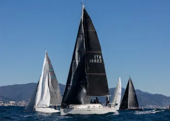Flotta di barche a vela con vele nere e bianche in regata su mare mosso, con coste collinari sullo sfondo e cielo sereno