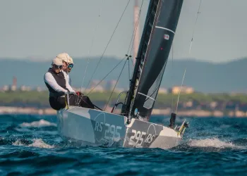 Tre velisti a bordo di una barca RS21 con numero velico 295 navigano con mare mosso, vela scura tesa e costa sullo sfondo.