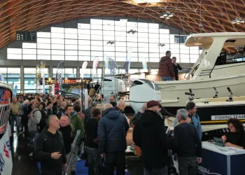 Visitatori affollano un padiglione fieristico tra barche e motori fuoribordo esposti, sotto una grande copertura in legno