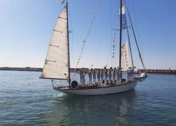 Veliero d'epoca con scafo bianco e vele spiegate in porto, con l'equipaggio in divisa schierato sul ponte che saluta alzando i cappelli.