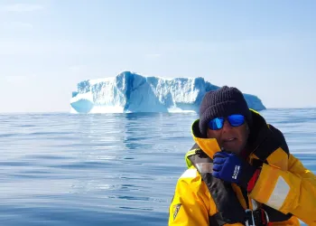 Persona con giacca gialla e occhiali da sole su una barca vicino a un grande iceberg in mare calmo.