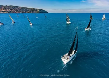 Veduta aerea di una flotta di barche a vela con vele nere che navigano su un mare blu, con la costa collinare sullo sfondo.
