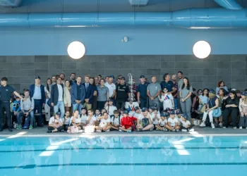 Gruppo di adulti e giovani atleti seduti e in piedi accanto a una piscina interna durante un evento.