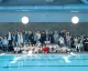Gruppo di adulti e giovani atleti seduti e in piedi accanto a una piscina interna durante un evento.