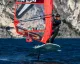 Windsurfer foil con vela rossa e numero ITA 294 su acqua davanti a una costa rocciosa.