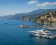 Yacht di lusso ormeggiati in un porto turistico costiero con borgo e scogliere sullo sfondo
