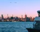 Skyline urbano al tramonto visto dall'acqua, con un traghetto in primo piano a destra.