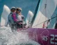 Equipaggio a bordo di un monotipo RS in regata con spruzzi d'acqua e vele sullo sfondo
