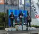 Tre uomini sul podio della premiazione davanti al lago, con bandiere e pannello blu sullo sfondo; quello al centro regge un trofeo