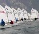 Una lunga fila di Optimist in linea di partenza sull'acqua mossa, con i velisti in assetto e le montagne sullo sfondo.