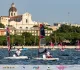 Barche OpenSkiff con vele rosse e nere in regata davanti al lungomare di Cagliari, con una chiesa a cupola sullo sfondo