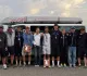 Gruppo di velisti e allenatori in posa davanti a un furgone con una barca ILCA sul tetto, alcuni con medaglie e targhe, sul lungomare